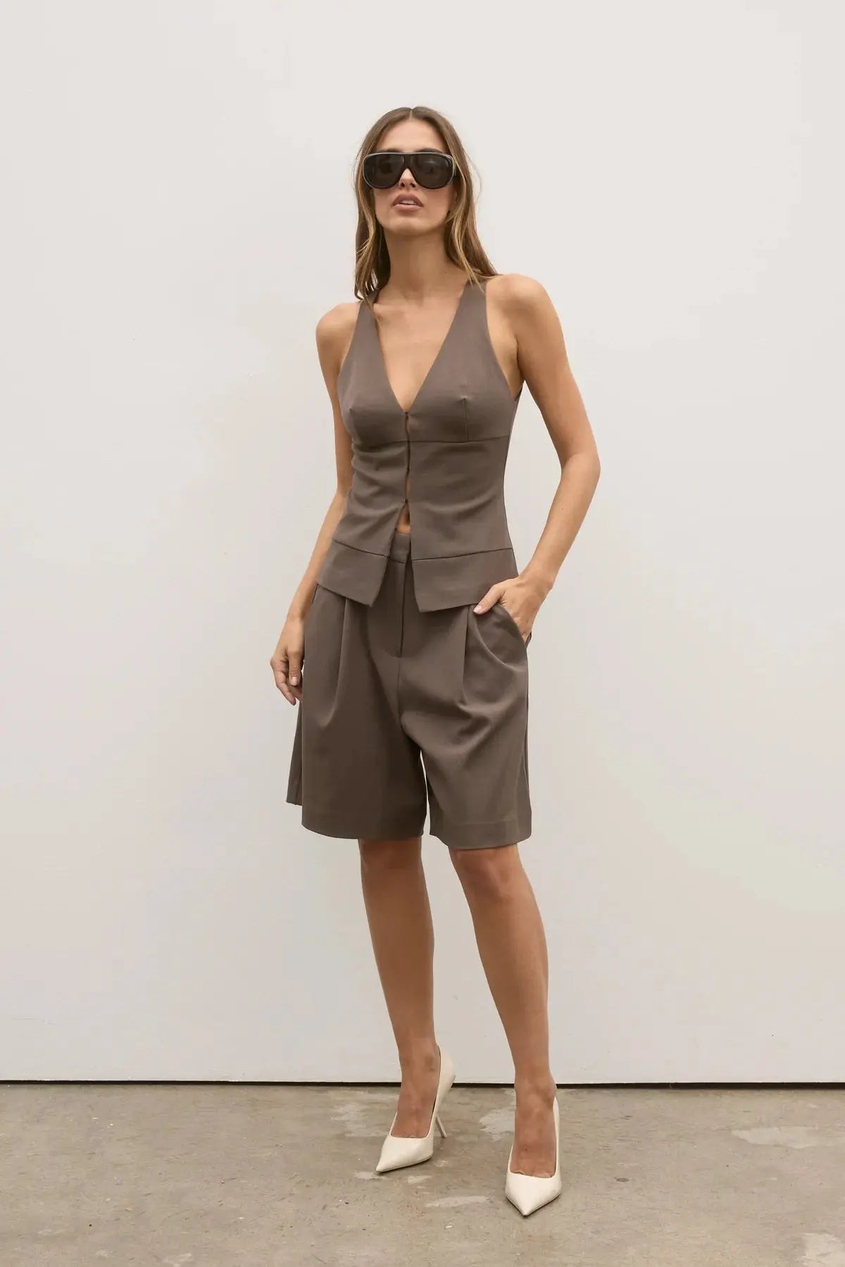 Woman wearing a brown sleeveless top and shorts set with sunglasses against a white background